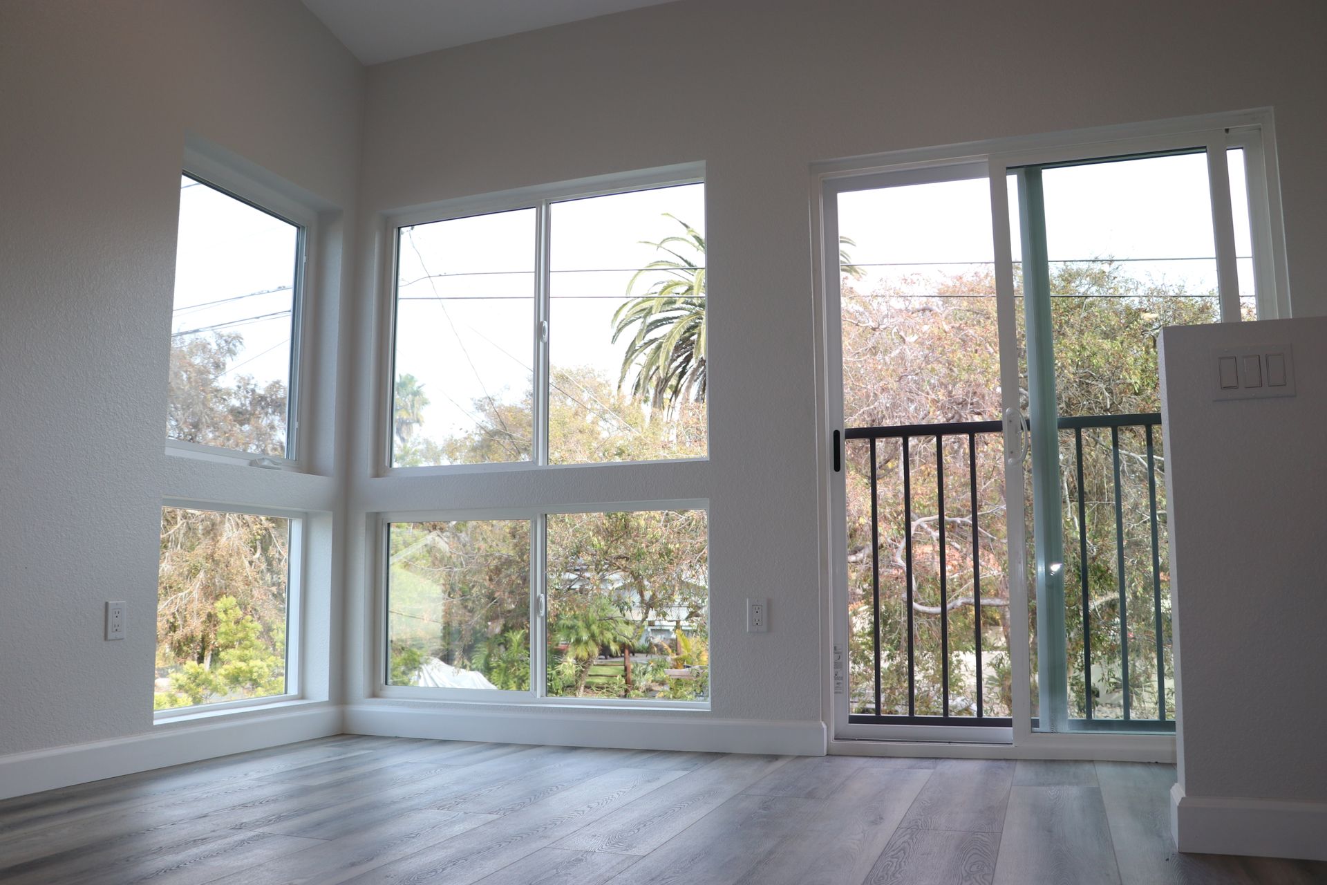 Bright empty room with corner picture windows and a sliding door opening to a railed balcony with treetop views.