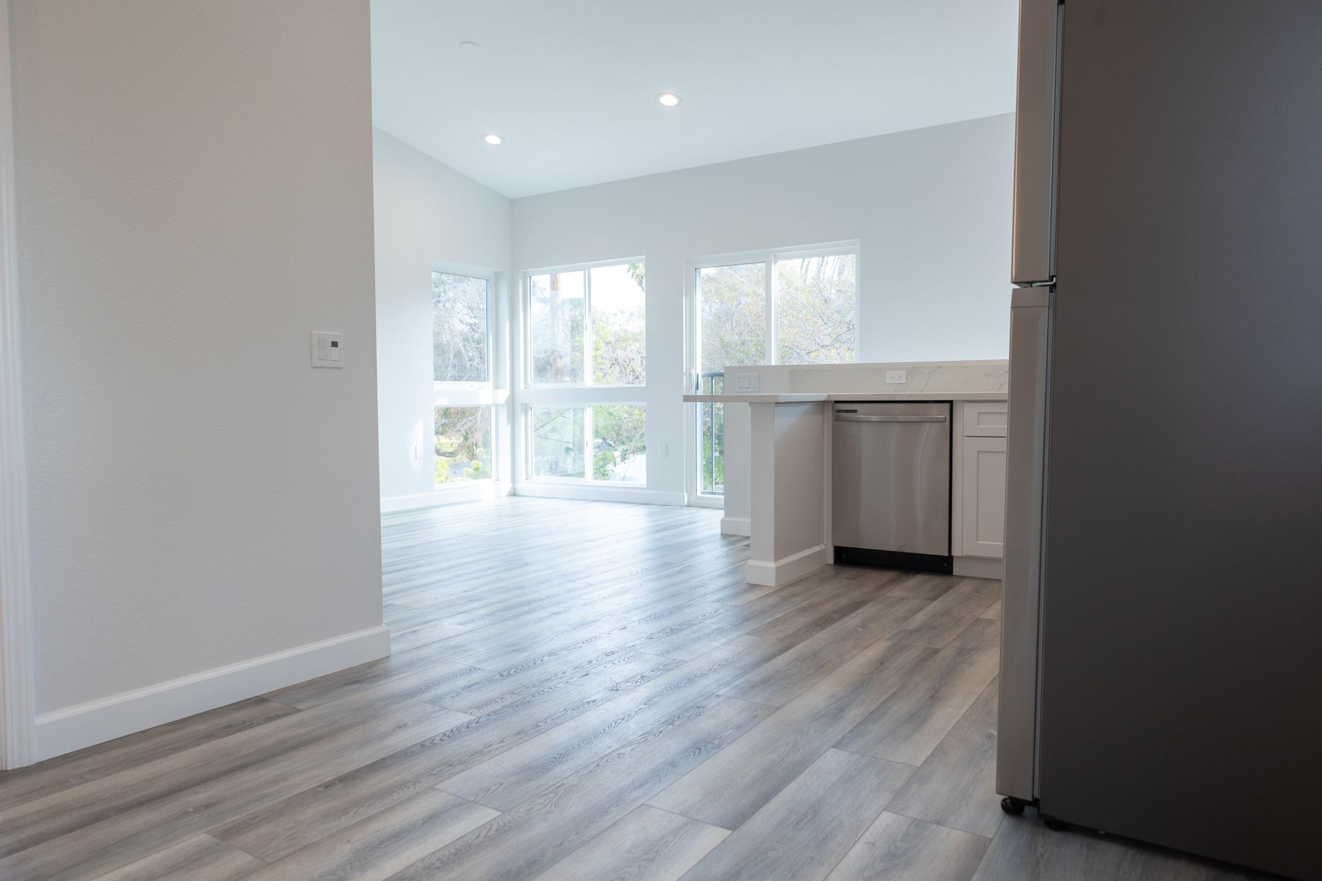 Empty ADU living area with corner windows, kitchen peninsula, and stainless dishwasher.