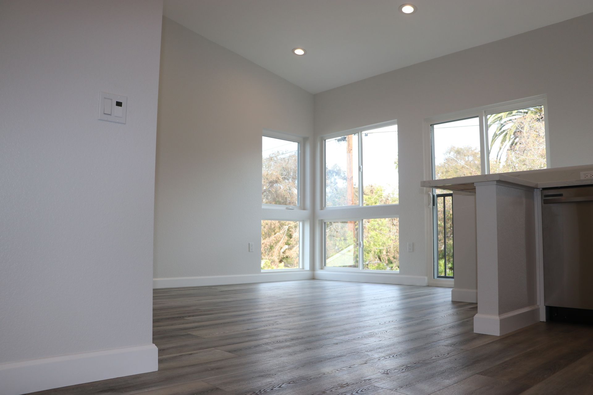 Empty living area with tall windows, recessed lighting, gray-wash vinyl plank flooring, and kitchen peninsula at right.
