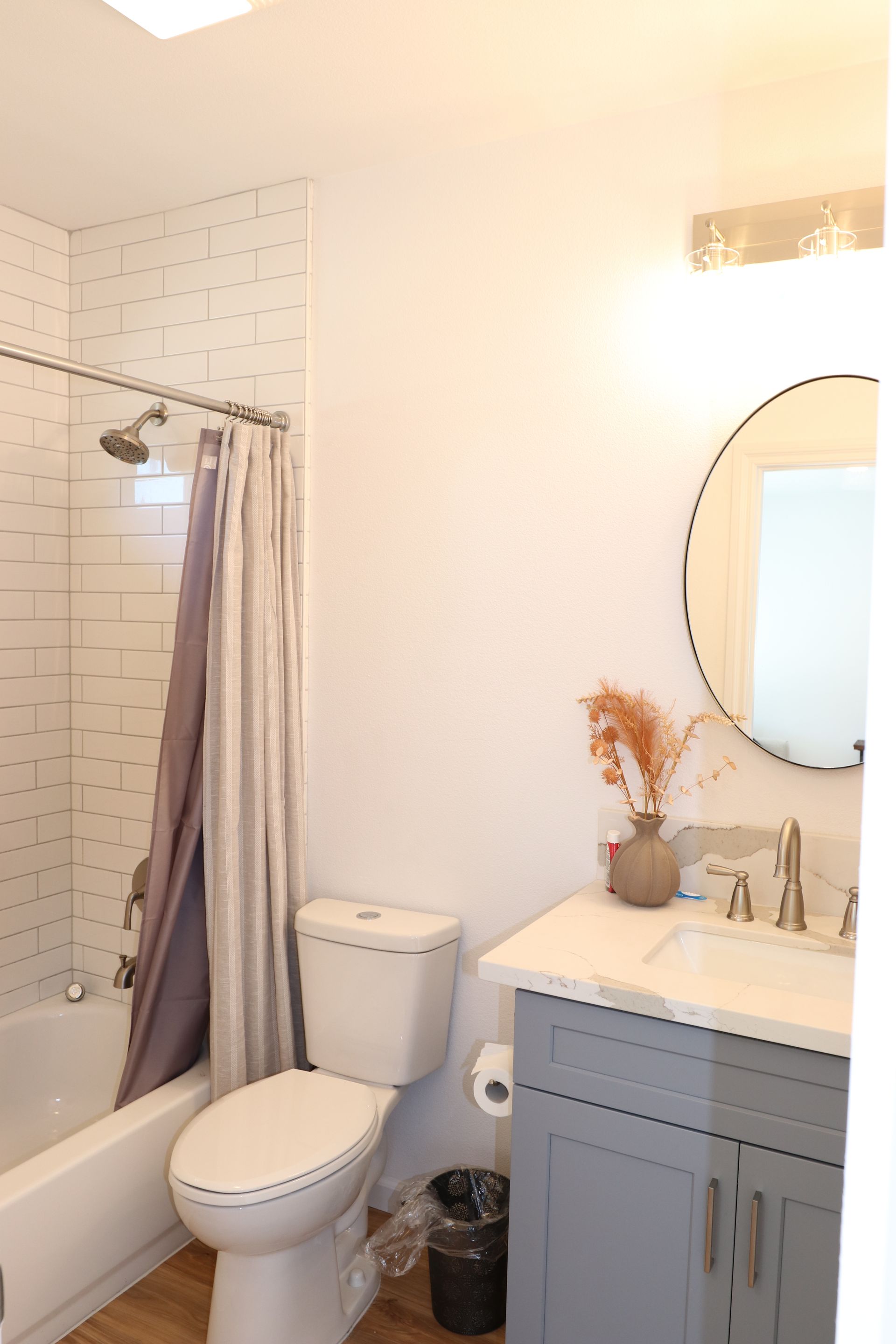 Bathroom with gray vanity, quartz counter, round mirror, and subway-tile tub shower with curtain.