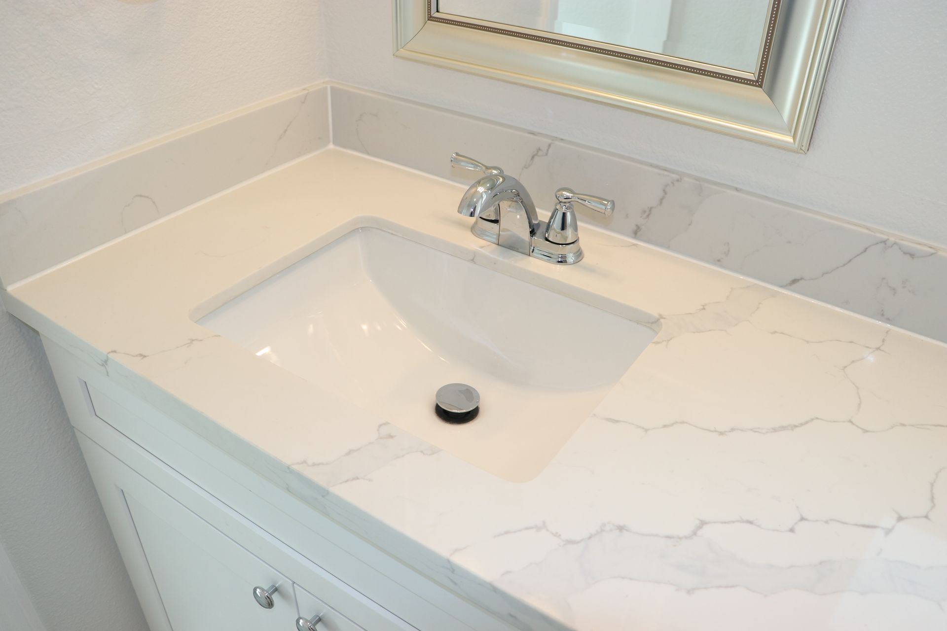 White bathroom vanity with marble-look quartz countertop, undermount sink, and chrome faucet below a framed mirror.