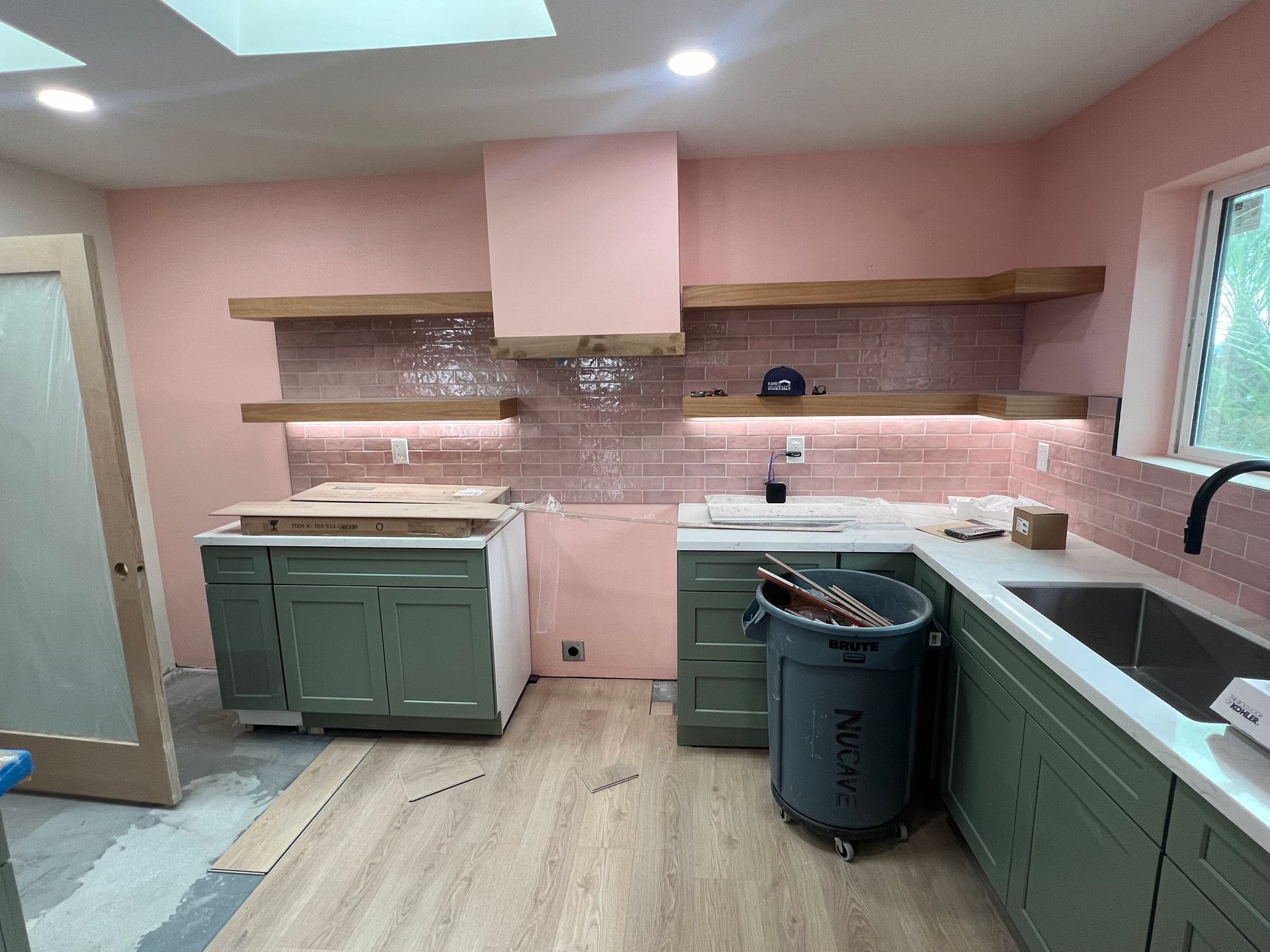 Kitchen nearing completion with sage green cabinets, pink tile backsplash, and finish work underway.