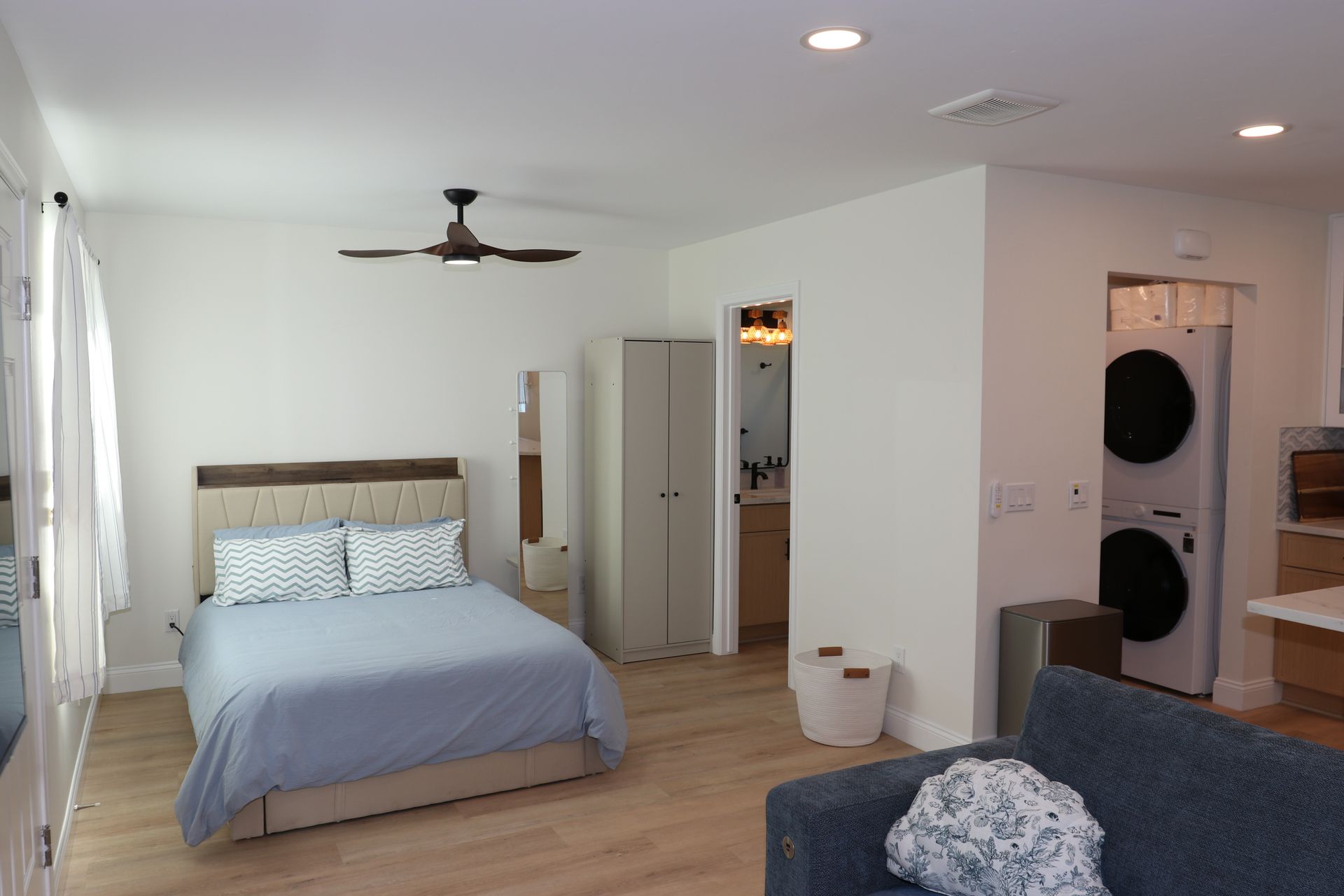 Studio ADU sleeping area with upholstered bed, ceiling fan, and stacked laundry closet beyond.